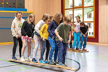 Teambuilding! Sechs Schülerinnen und Schüler stehen auf zwei Brettern, die sie wie Skier unter ihren Füßen haben, und Laufen gemeinsam durch eine Turnhalle. Nur wenn sie ihre rechten bzw. linken Beine gemeinsam heben, gelingt das gemeinsame Vorankommen.