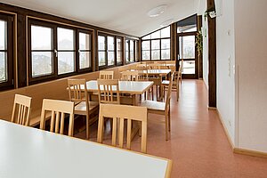 Blick in den Speisesaal. Unter der Fensterfront befindet sich an eine lange Bank. Tische mit Stühlen stehen hier nebeneinander aufgereiht. 
