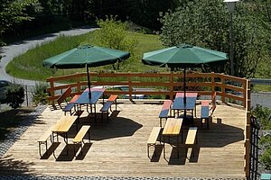 Blick auf die Holzterasse mit vier Biertischgarnituren und zwei Sonnenschirmen.