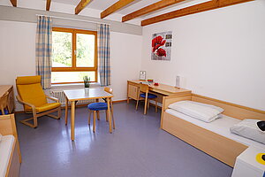 Blick ins Zimmer für eine Lehrkraft. Es ist ausgestattet mit Bett, Tisch uns Stuhl und einem Sessel am Fenster. 
