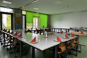 Im Speisesaal sind die Tische in Kreisform aufgestellt. An den Fenstern hängen grüne Vorhänge.