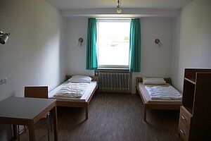 Blick in ein Zweibettzimmer. Die Betten stehen links und rechts vom Fenster. Außerdem befinden sich ein Tisch mit Stuhl und ein offener Schrank im Zimmer. 