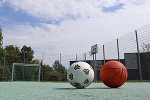 Ein Fußball und ein Basketball liegen auf der grünen Fläche des Sportplatzes.
