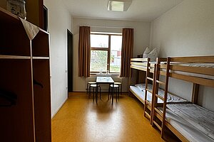 Ein Mehrbettzimmer mit Stockbetten auf der einen Seite des Zimmers und Regalen zur Ablage des Gepäcks auf der anderen Seite. Am Raum Ende befindet sich das Fenster.