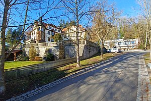 Blick auf das Obersteinbacher Schloss von der Auffahrt aus. Auf der linken Seite blickt man über eine Mauer in den kleinen Park, der zum Gelände gehört. Das Schloss steht leicht versteckt hinter herbstlich entlaubten Bäumen. 