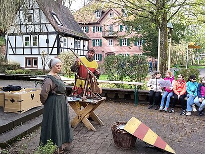 ein Mann und eine Frau in mittelalterlichen Gewändern erzählen Kindern auf einem Platz vor Fachwerkhäusern