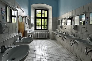 Blick in einen Waschraum mit vielen Waschbecken an den gefließten Wänden. Am Ende des Raums befindet sich ein hohes Sprossenfenster. 