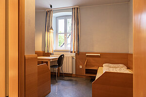 Der Blick in ein Zimmer für Lehrkräfte und Betreuungspersonen. Es sind ein Tisch mit Stuhl und ein Bett zu sehen. 