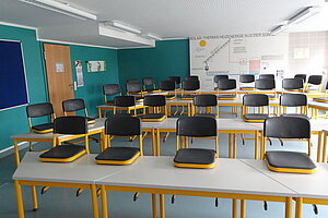 Ein Klassenhimmer, in dem die Tische frontal ausgerichtet sind. Stühle sind auf die Tische geräumt.