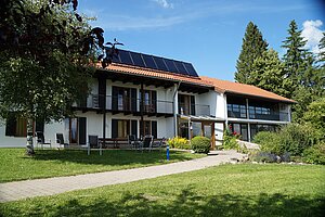 Zweistöckiges Haus mit umlaufenden Balkonen, Solaranlage auf dem Dach und grünem Umfeld am Hang.