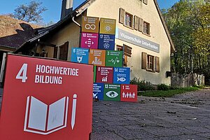 Vor dem Haus in dem sich die Umweltstation befindet stehen Würfel auf denen die Ziele der Nachhaltigen Entwicklung abgebildet sind. Im Vordergrund ist das Ziel Nummer 4 "Hochwertige Bildung" zu sehen. 