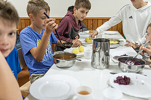 Schüler beim Mittagessen am Tisch. Das Essen wird aus Schüsseln auf die Teller verteilt. 