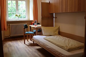 Blick in ein Zimmer mit Bett, Tisch und Stuhl. Am Fenster hängen gelbe Stoffvorhänge.