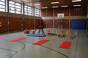 Blick in die Sporthalle, mit aufgereihten Turngeräten (Bock, Sprungbrett, Kästen), Matten und Hütchen.